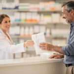 Pharmacist handing a prescription bag to a smiling customer at the pharmacy counter.