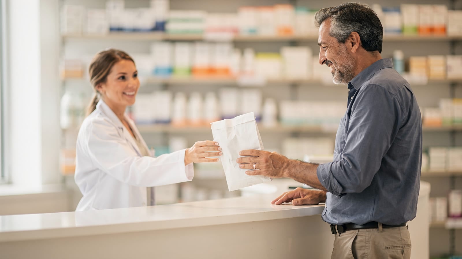 Pharmacist handing a prescription bag to a smiling customer at the pharmacy counter.